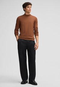 Mann mit lockigem Haar in einem braunen Rollkragenpullover und schwarzen Hosen steht selbstbewusst und schaut zur Seite vor einem neutralen Hintergrund.