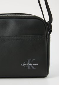 Black synthetic handbag with a smooth texture, featuring a zipper and a front pocket. Engraved “Calvin Klein Jeans” logo in white.