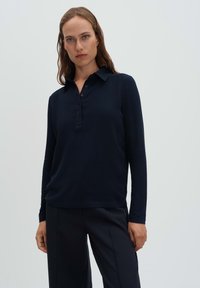 Donna che indossa una polo a maniche lunghe blu scuro con bottoni e pantaloni abbinati blu navy contro uno sfondo chiaro e semplice.
