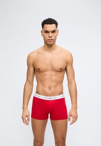 Oberkörperfreier Mann mit lockigem Haar, der rote Tommy Hilfiger Boxershorts trägt, vor schlichtem hellgrauem Hintergrund stehend.