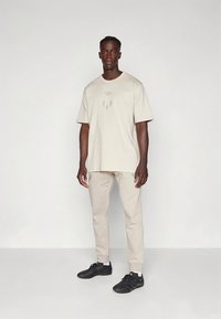 Μπεζ φαρδιά μπλούζα με διακριτικό λογότυπο, συνδυασμένη με μπεζ στενά παντελόνια και μαύρα παπούτσια, με απαλό ύφασμα και άνετη εφαρμογή.