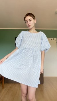 Personne aux cheveux courts portant une robe à carreaux bleu clair et blanc, tenant la jupe, debout dans une pièce aux murs verts et au sol en bois.