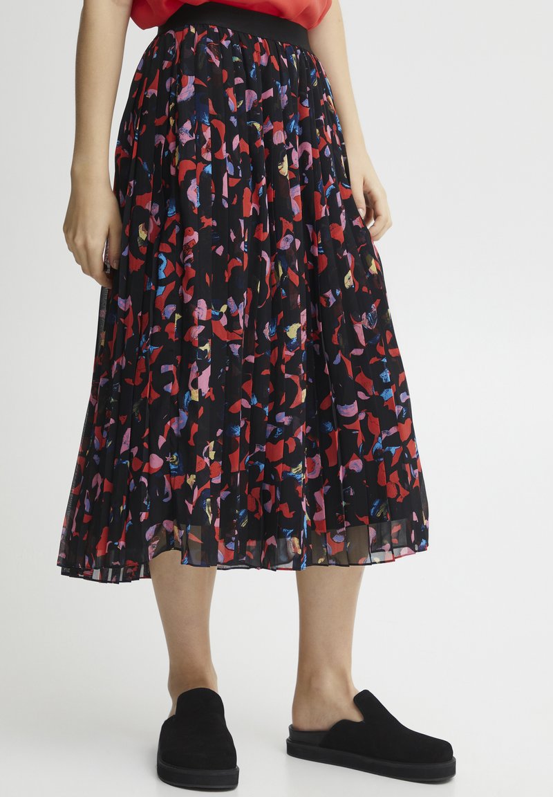 Een zwarte geplooide rok met een kleurrijk abstract patroon in rood, roze en blauw. De tailleband is zwart, gecombineerd met zwarte instapschoenen.