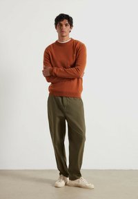 Pomarańczowy sweter dziany z okrągłym dekoltem, w połączeniu z luźnymi oliwkowymi spodniami i beżowymi sneakersami, stojący na jednolitym tle.