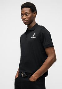 Νέος άνδρας που φοράει μαύρο πόλο με λογότυπο φοίνικα και κείμενο στο στήθος, με τα χέρια στις τσέπες, στέκεται μπροστά σε απλό φωτεινό φόντο.