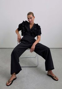 Outfit nero con una blusa a maniche corte arricciata e pantaloni a gamba larga. Indossato con sandaletti neri piatti. Sfondo e sedia grigi.