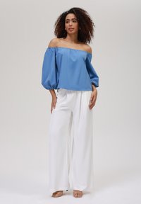 Blå av-shoulder bluse med puffede ærmer, parret med hvide brede bukser og lysebrune sandaler med stropper. Glat stoftekstur.