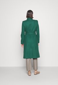 Groene wollen blend jas met een getailleerde pasvorm, lange mouwen, een ceintuur in de taille en een achterste split, gecombineerd met beige broek en lichte schoenen.