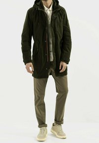 Hombre con parkas con capucha verde oscuro, camiseta polo blanca, pantalones caqui con puños y zapatillas beige, de pie contra un fondo blanco.