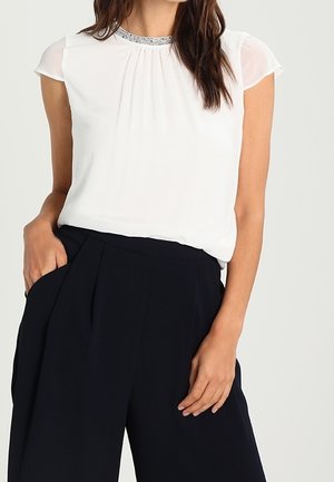 Vrouw draagt een witte mouwloze blouse met een kralenversierde halslijn en zwarte, hoge taille, wijde broekspijpen, één hand in de zak.