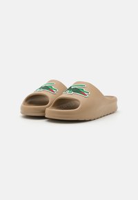 Bruine instapsandalen van soepel materiaal, met een afgeronde open teen, textuurzool en groene geborduurde logo-accenten.