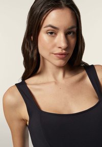 Musta ilmanhiha toppi, jossa on neliömäinen pääntie. Sujuva kangas, jossa on saumat edessä ja vartalonmyötäinen muotoilu, joka näyttää minimalistiselta.