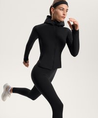 Černá sportovní bunda s vysokým límcem a plným zipem, v kombinaci s černými legínami a lehkými běžeckými botami. Hladká textura po celém povrchu.