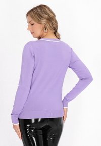 Maglione lungo viola a maniche lunghe con collo rotondo, dettaglio bianco al colletto e polsini a costine, abbinato a pantaloni neri lucidi.