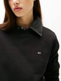 Svart sweatshirt med rund hals og denimkrage. Har et lite brodert logo i rødt, hvitt og blått på brystet.