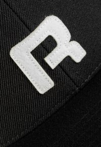 Schwarze Mütze mit einem auffälligen weißen gestickten Logo. Der Stoff erscheint strukturiert mit sichtbaren Nähdetails.