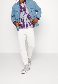 Veste en jean bleu clair, chemise à motifs violets et pastel, pantalons blancs et baskets montantes grises. Tenue décontractée en couches.