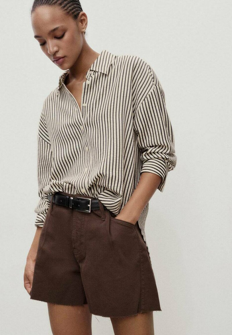 Chemise boutonnée à rayures beiges et noires rentrée dans un short marron, avec des poches et un ourlet effiloché, maintenue par une ceinture noire.
