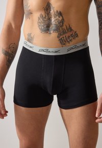 Fekete pamut boxer alsónadrág, szürke elasztikus derékpánttal, kézzel írt logóval. Testhez simuló fazon, sima textúrával és varrásokkal.