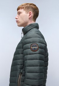 Donkergroen pufferjack met gestikte padding, hoge kraag en een patch op de mouw. Voorzien van een ritszak met contrasterende rits.