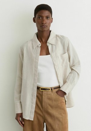 Femeie purtând o cămașă bej cu nasturi peste un maiou alb și pantaloni maro din piele întoarsă, cu o centură, stând în fața unui perete simplu și luminos.