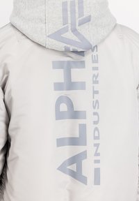 Světle šedá zateplená bunda s kontrastní šedou kapucí. Na zadní straně se nachází velký textový grafický nápis "ALPHA INDUSTRIES". Hladká textura.