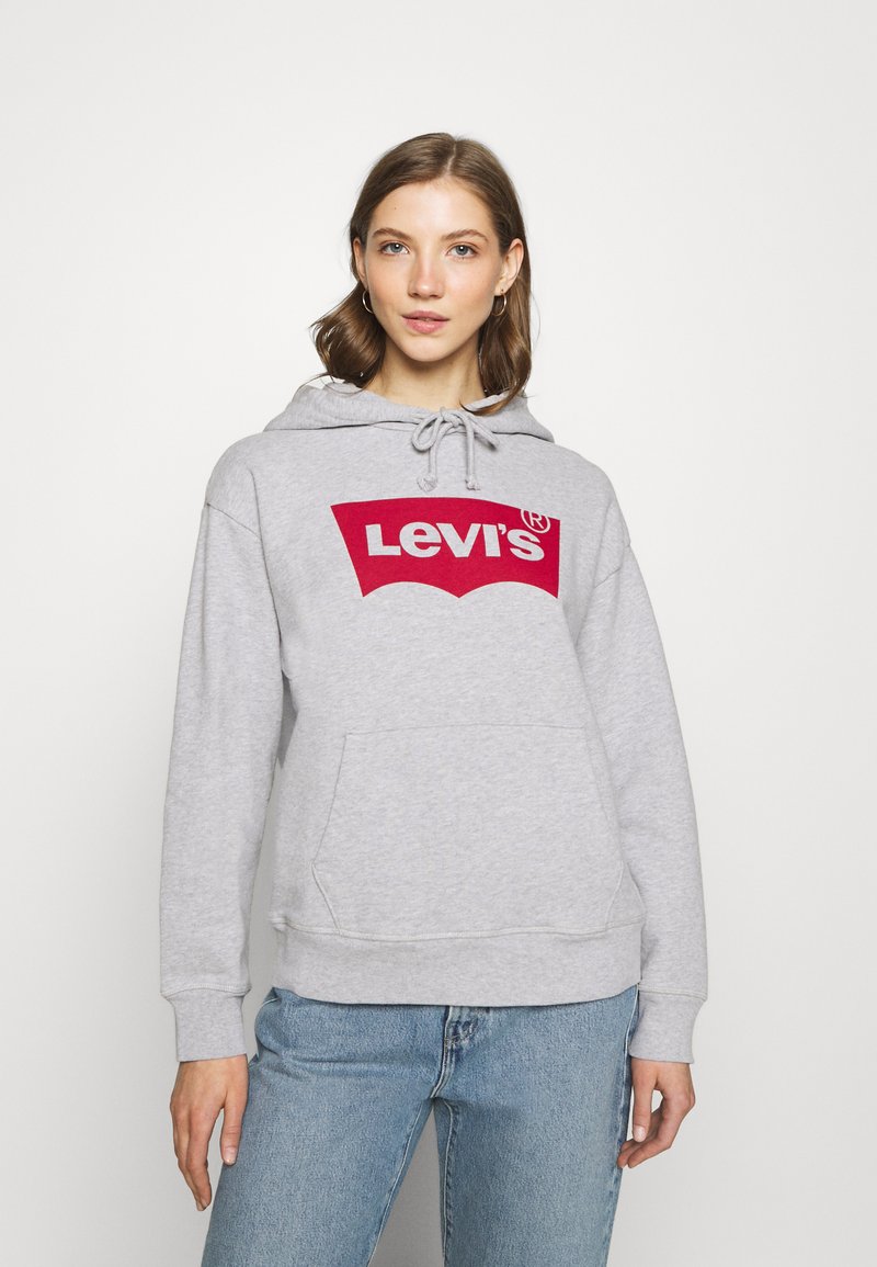 Felpa grigia con cappuccio, caratterizzata da un grande logo rosso di Levi's, cappuccio con coulisse, tasca anteriore e polsini a costine. Indossata con jeans azzurri chiari.