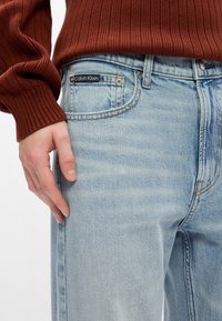 Gaiši zili džinsu bikses ar izbalējušu dizainu, vara šuvēm, priekšējo kabatu un melnu Calvin Klein logo uz jostas vietas.