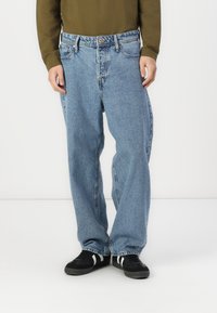 Hellblaue Denim-Jeans mit lockerer Passform, ausgestattet mit einem Fünf-Taschen-Design und Metallknopfverschluss. Tragen Sie dazu ein langärmliges olivgrünes Hemd.