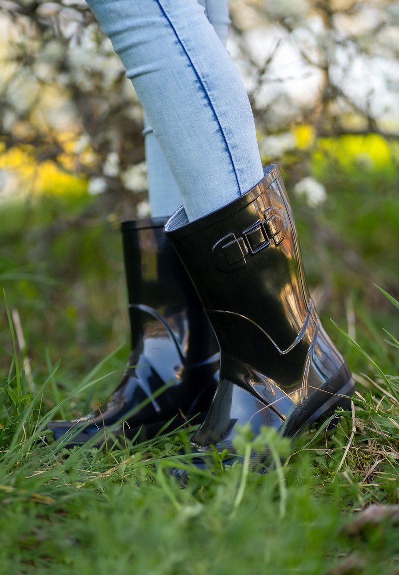 Bottines en caoutchouc noir étanches avec une finition brillante, dotées d'une sangle décorative et d'un détail de boucle, présentées sur une surface herbeuse.