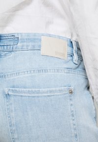Ljust blå denimjeans med en rektangulär vit etikett med "DRYKORN" präglat, bakfickor och silverfärgat hårdvara vid midjan.