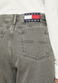 Vaqueros grises con una etiqueta de Tommy Jeans en la cintura. El atuendo se complementa con una parte superior blanca parcialmente visible por encima de la cintura.