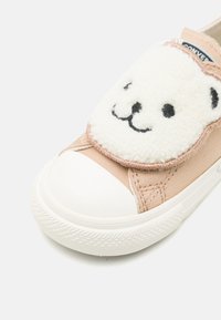 Zapatilla de lona beige con un parche de cara de oso peludo blanco en la correa de Velcro y puntera de goma, con suela texturizada y detalles cosidos.