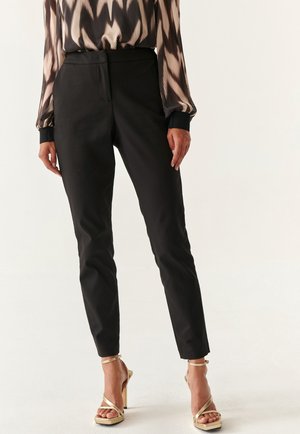 Black tailored trousers with a slim fit, smooth texture, and high waist. Paired with gold strappy high-heeled sandals.