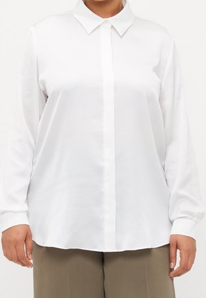 Persona vistiendo una camisa blanca de manga larga y botones, de corte holgado, con cuello puntiagudo y pantalones verde oliva.