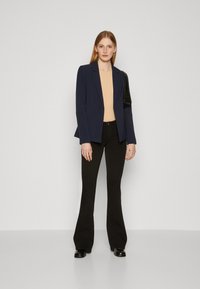 Navy blazer with a tailored fit, beige top, and black flared trousers; model holding a black handbag, minimalistic design, smooth textures.