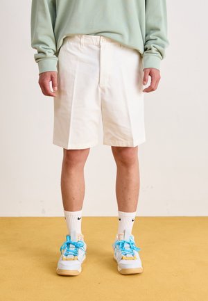 Person iført hvide knælange shorts, lyseblå sweatshirt, hvide Nike sokker og hvide sneakers med blå snørebånd, der står på gul gulv.