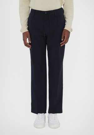 Pantaloni blu navy con vestibilità dritta, dotati di vita con coulisse, tasche laterali e una texture del tessuto liscia. Indossati con sneakers bianche.