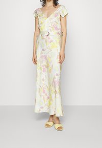 Maxi-jurk met bloemenprint in pastelkleuren (geel, roze, blauw) met een V-hals, korte mouwen en een taille met ceintuur. Gecombineerd met gele sandalen.
