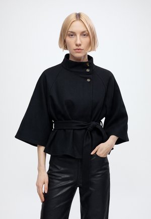 Femme aux cheveux courts et blonds portant une veste noire ceinturée aux manches larges et un pantalon en cuir noir, debout sur un fond blanc.