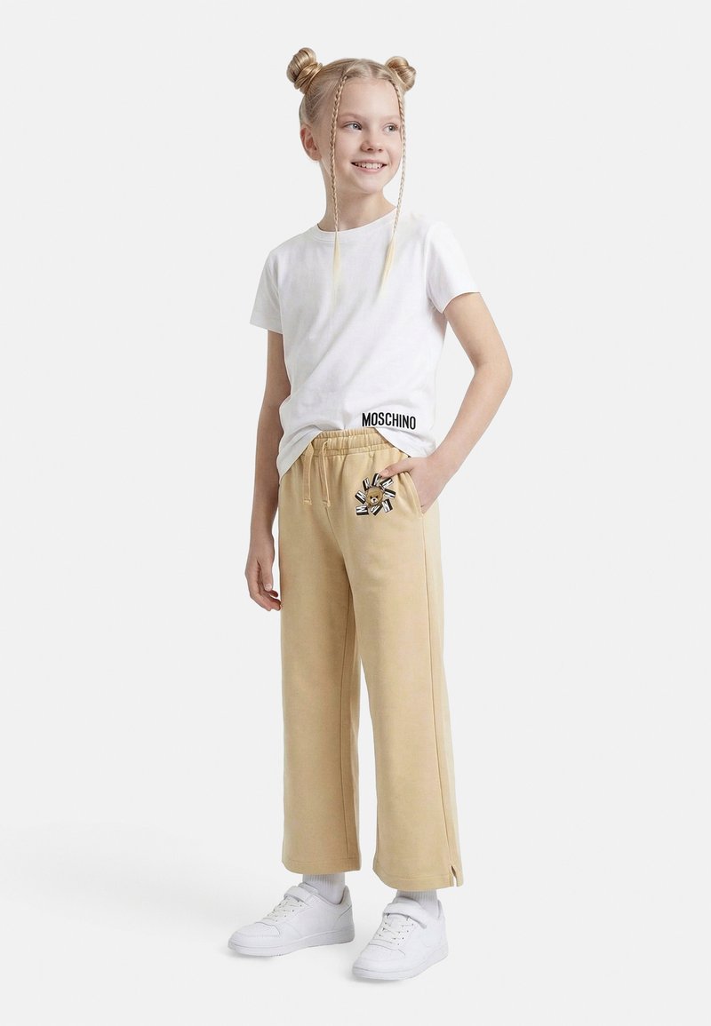 Χαμογελαστή κοπέλα με ξανθά μαλλιά σε διπλούς κότσους και πλεξούδες φοράει λευκό μπλουζάκι Moschino, μπεζ παντελόνι φαρδύ με στάμπα αρκουδάκι και λευκά αθλητικά παπούτσια.