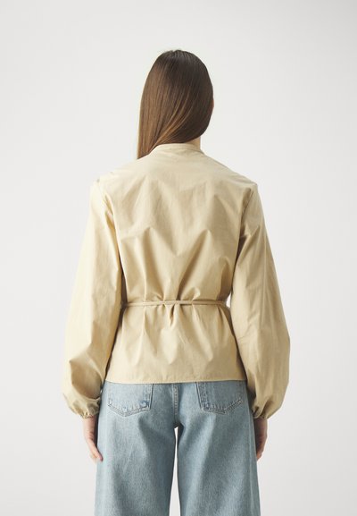 Beige getailleerde jas met een cinched taille, lange pofmouwen en een hoge halslijn, gecombineerd met lichtblauwe denim jeans met wijde pijpen.