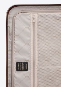 Interno della valigia rigida marrone con rivestimento beige, tasca in rete, compartimenti con cerniera e etichetta con logo. Superficie testurizzata, layout organizzato.
