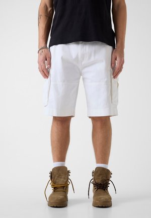 Mężczyzna ubrany w białe szorty bojówki, brązowe buty sznurowane z różnokolorowymi sznurówkami oraz czarną koszulę, stojący z rękami po bokach.