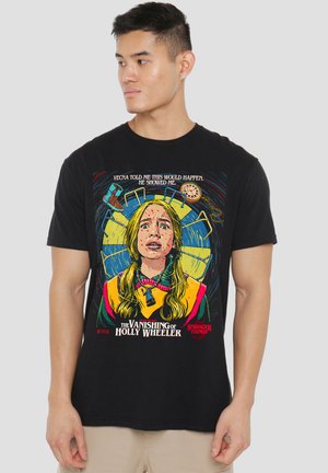 Man wearing black Stranger Things t-shirt featuring distressed girl with pigtails, playground equipment, and text about “The Vanishing of Holly Wheeler.”