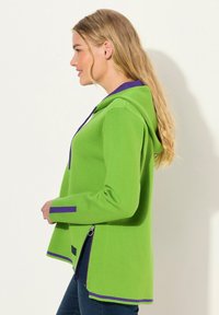 Giacca verde con cappuccio e accenti viola, presenta una zip ai lati, una vestibilità rilassata e una texture liscia.