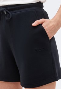 Άτομο που φοράει μαύρα σορτς Borg με το χέρι τοποθετημένο στην μπροστινή τσέπη, παρουσιάζοντας ελαστική μέση με κορδόνι και διακριτική λεπτομέρεια λογότυπου.