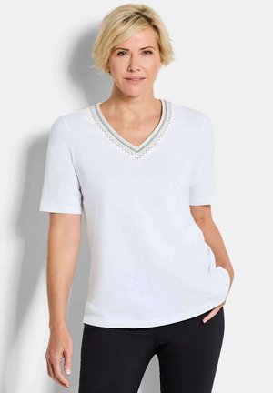 Frau mit kurzem blondem Haar, trägt ein weißes V-Ausschnitt-Shirt mit dekorativem Besatz, schwarze Hose, steht mit einer Hand in der Tasche.