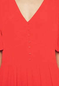 Rode jurk met V-hals en korte mouwen. Heeft een rij van zes knopen aan de voorkant en een zwierige rok met een geplooide textuur.