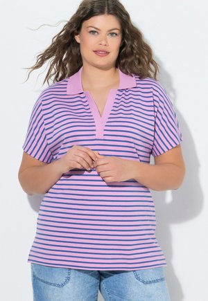 Femme aux cheveux bruns ondulés porte un polo à manches courtes rayé rose et bleu et un jean bleu clair, debout devant un fond uni.
